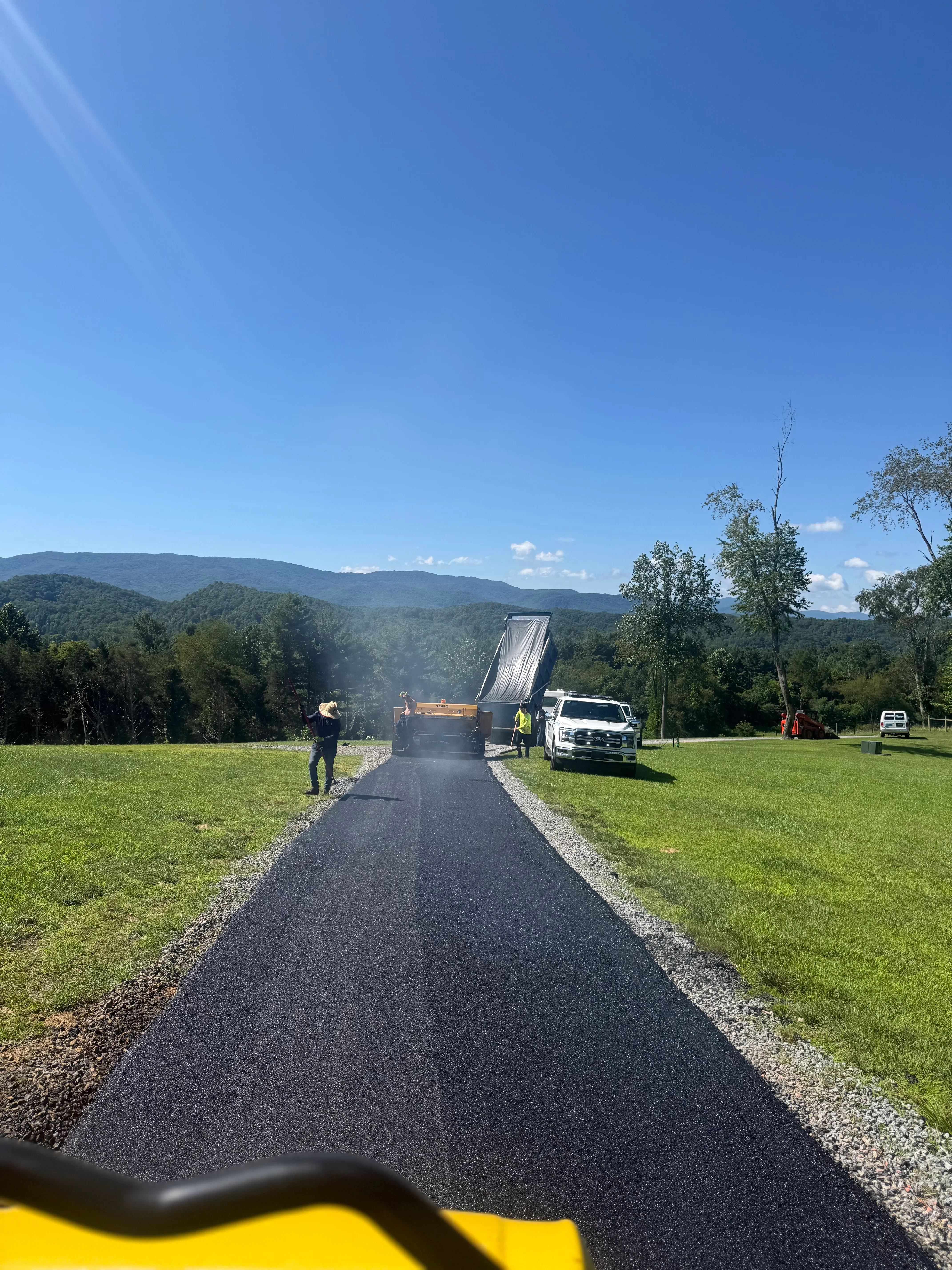 Newly installed black asphalt driveway with gravel borders and paving truck on residential property
